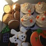  Photo shows a bakery box full of autumn treats: peanut butter iced chocolate cupcakes, oatmeal whoopie pies, Halloween themed royal iced sugar cookies. 